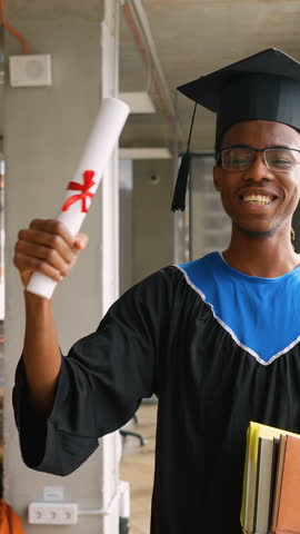African American graduate in cap and gown proudly holds diploma and books while standing in a bright modern academic space