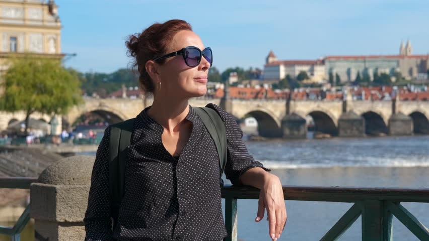 Tourist enjoying the view of charles bridge in prague. Woman traveler wearing sunglasses leaning against railing, overlooking iconic charles bridge spanning vltava river during bright day in prague