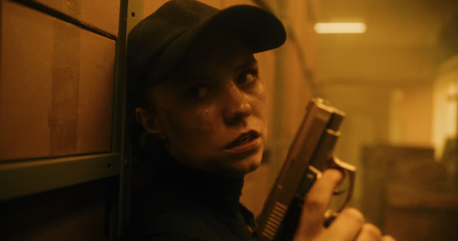 Close Up of Female Secret Agent in Tactical Gear Hiding Behind Cover, Holding a Pistol, Talking to Killer. Officer Preparing to Attack Criminal in Dark Warehouse. Epic Action Movie Scene. Warm Look.