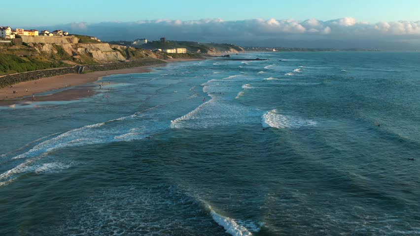 Scenic aerial drone shot of Biarritz coastline, with surfers in Atlantic waves, sandy beach, and cliffs under partly cloudy sky. Popular French surf spot, France