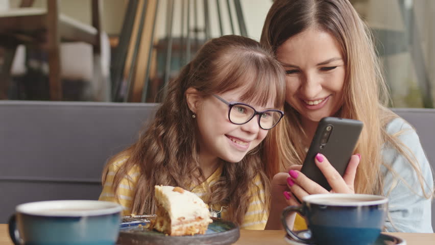 Waist-up slowmo of joyful young woman and her 11-year-old daughter with down syndrome looking at photos on smartphone and laughing while sitting at table in cozy cafe