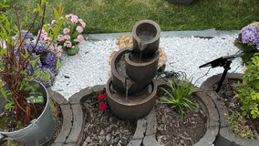 A modern tiered garden fountain flows gently among blooming flowers and mulch, powered by a visible solar panel. White stones and lush greenery complete this tranquil outdoor setup. - Powered by Shutterstock - Get 15% off with code: PIKWIZARD15