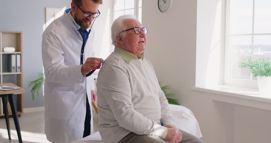Mature senior male patient sitting on couch in clinic for a medical checkup. Physician doctor with stethoscope examining man's lungs, breathing and heartbeat at the hospital in exam room. 4k video.