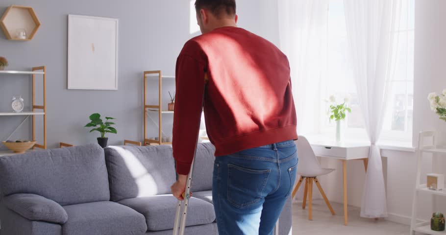 Man with trauma, broken leg, man wearing foot support with walking stick crutches, patient wearing sling support after accident, traumatic stress, accident, sitting at home on sofa, resting to relax