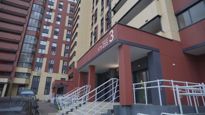 Entrance of modern building located in courtyard next to parked cars. High-rise within residential complex features dark and light facade