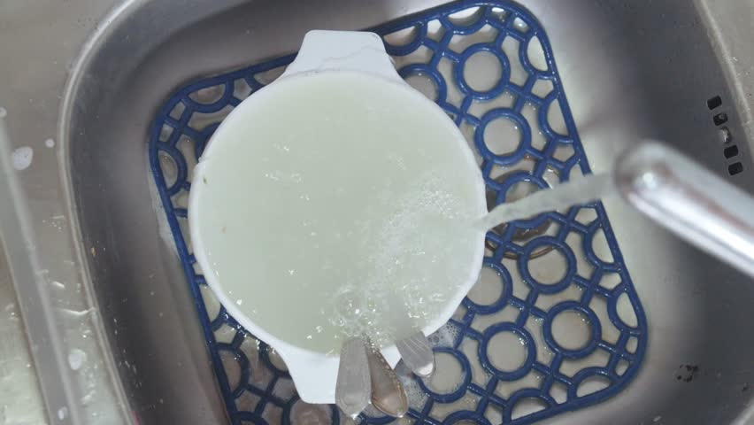 Water flows from a faucet into a sink with dirty dishes covered in suds from detergent.