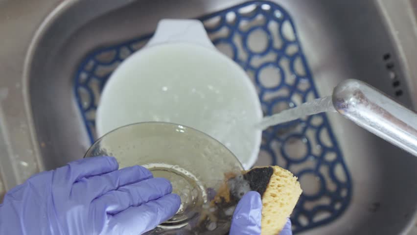 A hand in a blue glove washes a plate in a sink under a stream of water with suds from detergent.
