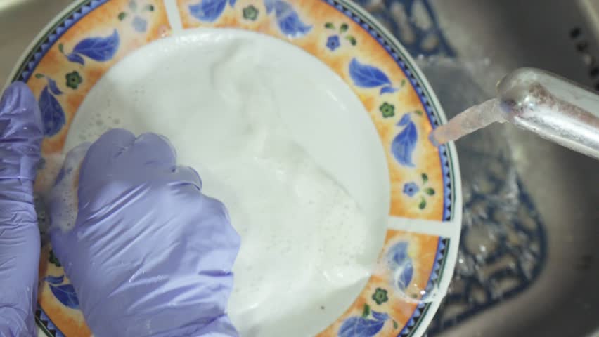 A gloved hand washes dirty dishes with a sponge in a sink under running water with suds.