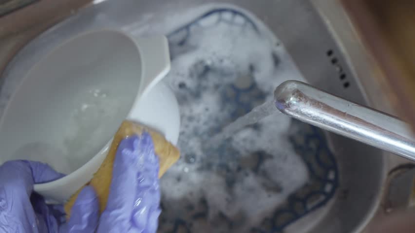 A hand in a blue glove washes a bowl with a sponge under running water in a sink with suds.