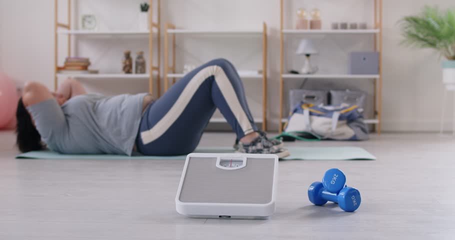 Close up shot of scales and dumbbells with overweight girl doing abdominal crunches on background. Young plus size fat woman doing sport exercises lying on yoga mat. Workout sport and slimming concept