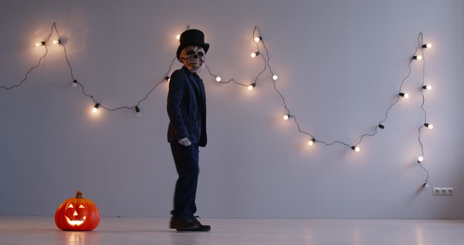 Happy child boy in Halloween black dress, wearing a scary rubber mask and hat taking big wide steps and having fun near the glowing jack-o-lantern on the background of a garland of light bulbs. 