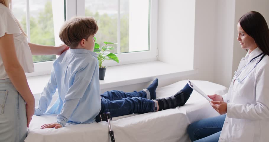 Female traumatologist talking about treatment and examining little kid patient with injured leg sitting on hospital bed. Boy in orthopedic bandage is supported by his mother during medical inspection.