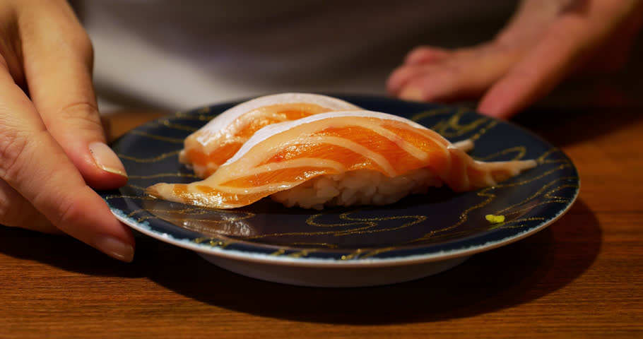 Close-up of fresh salmon belly nigiri on black plate, woman turning plate to display thick marbled cuts from both sides, showcasing premium quality and rich texture. Casual sushi conveyor restaurant.