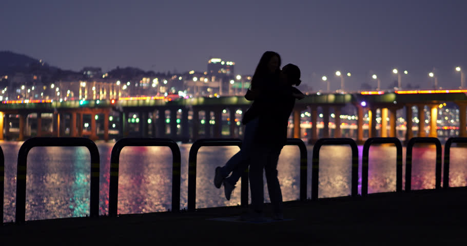 Silhouetted couple embracing joyfully at riverside park by Han River, man lifts woman and spins around as they laugh together. Blurred background, city lights and illuminated Hannam Bridge glow behind