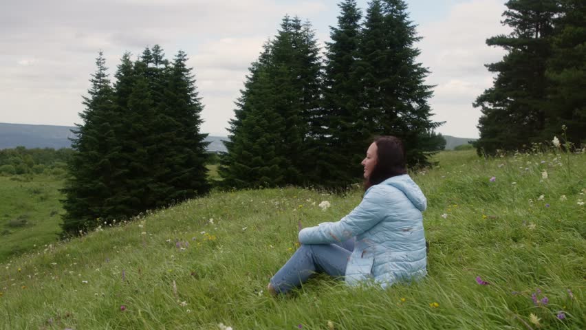 Young woman hiker sitting on a grassy hill, enjoying the breathtaking landscape filled with vibrant green trees and colorful wildflowers during a serene spring day