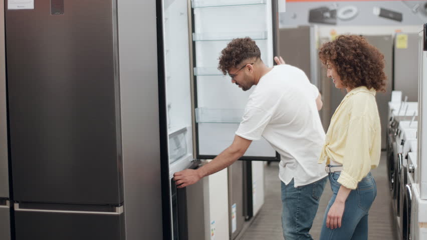 Couple browsing refrigerator models in appliance store, carefully examining potential purchase by touching surfaces, checking interior, evaluating design and features in slow motion
