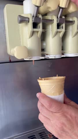 Close-up of chocolate soft serve ice cream being dispensed into a cone from a machine, hand holding cone, creamy texture and smooth swirl in real time