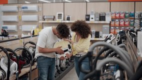 Diverse couple comparing robotic vacuum cleaner features, carefully analyzing product details within bright retail electronics store setting - Powered by Shutterstock - Get 15% off with code: PIKWIZARD15