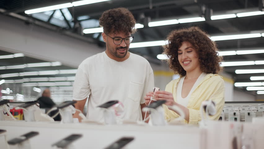 Young couple choosing charger for smartphone in electronics store. Customers browsing electronic store displays, carefully selecting smartphone accessories, comparing chargers.
