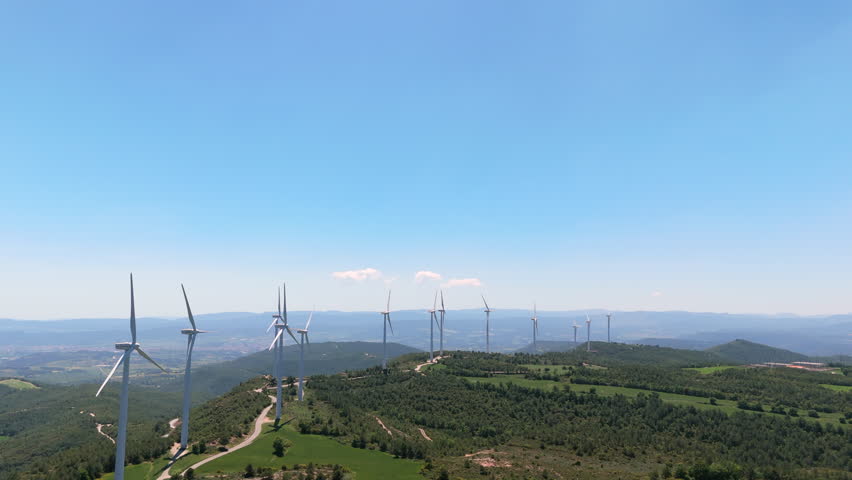 Aerial clip flying parallel to a row of wind turbines spinning on forested hills. Clear sky, clean energy and scenic mountain landscape captured by drone