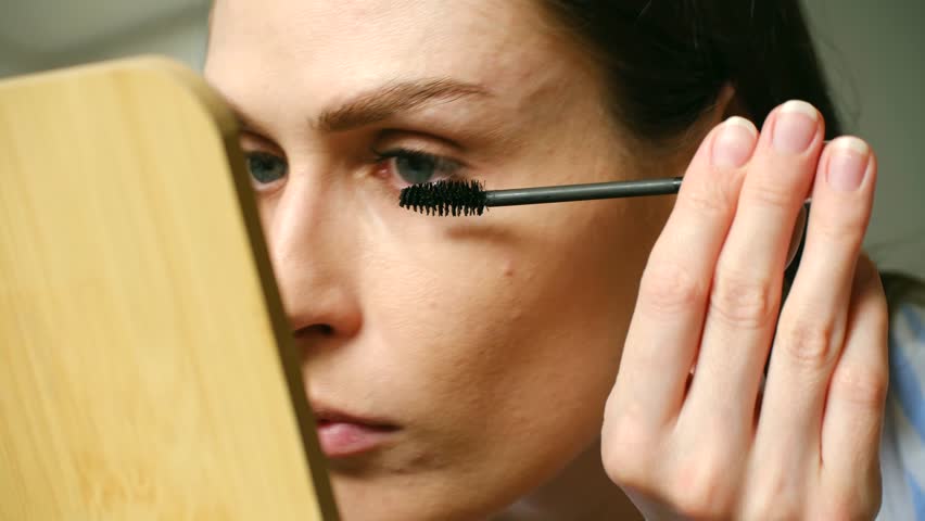 Caucasian woman carefully applies black mascara to her eyelashes, enhancing her eyes while looking in mirror during her daily makeup routine. Woman applying makeup mascara on eyelashes in front mirror