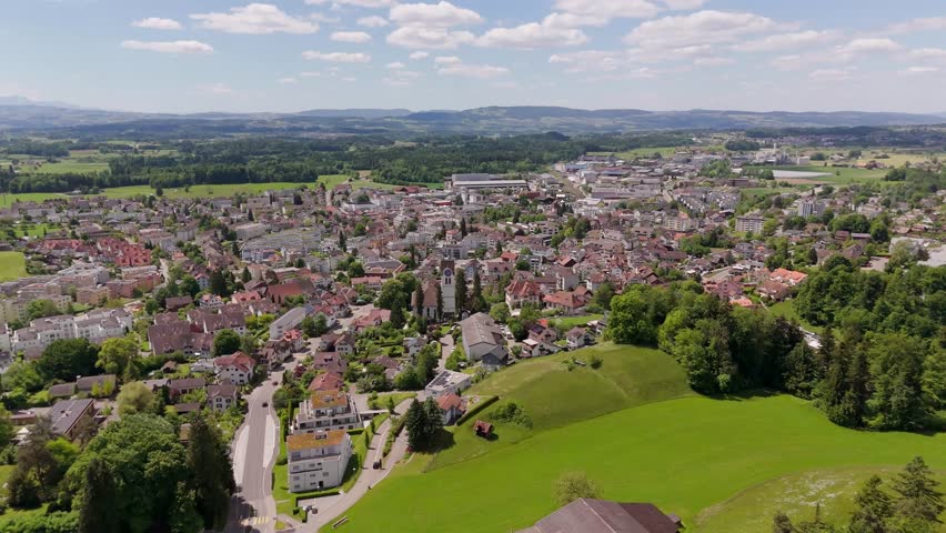Aerial flyover historic town of Hinwil in Switzerland. Church tower and houses in center of city. Wide shot. Sunny day with green picturesque landscape in distance. Clouds at sky in spring.