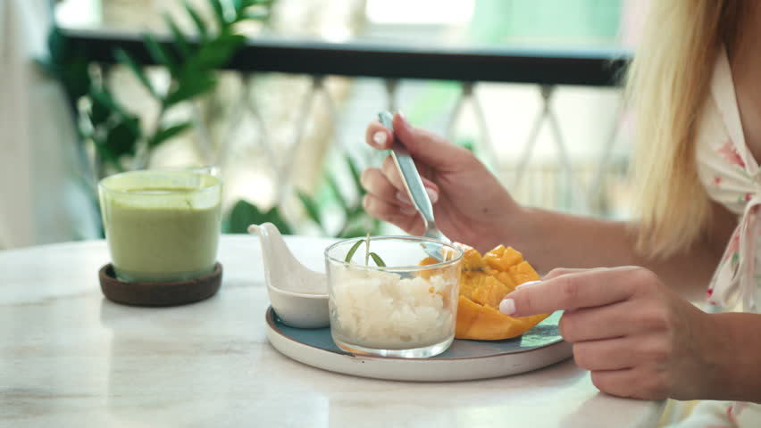 Close-up of woman enjoying traditional Thai mango sticky rice with coconut milk and matcha latte. Bright daylight cafe setting with tropical and fresh vibes.