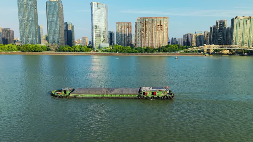 Aerial view of barge navigates river in urban setting with modern skyscrapers during clear day. Hangzhou, China.