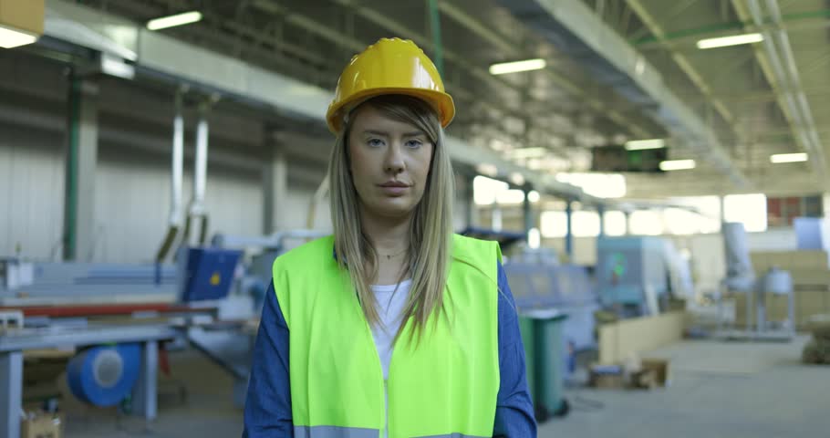 A young woman factory worker removes her yellow hard hat and smiles at the camera. She wears a denim shirt and green safety vest.