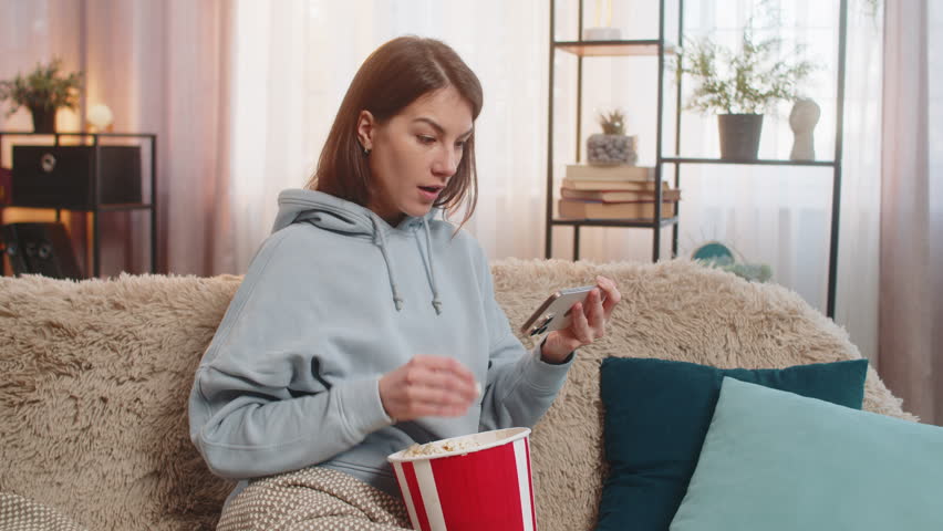 Happy Caucasian relaxed woman sits at home sofa watching movie film on smartphone eating popcorn. Young lady in casual clothes covered with blanket happily watching a series in living room apartment.