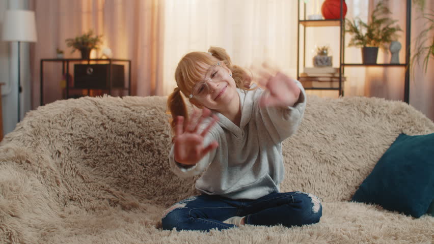 Young teenager girl waving hand with happy smile at home, expressing joy and friendliness. Caucasian child kid sitting on a sofa, saying hello hi welcome or goodbye with cheerful and relaxed attitude