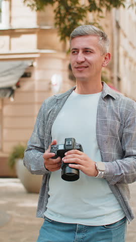 Middle-aged man tourist traveling on weekends exploring town looking around city, making photo pictures on camera outdoors. Caucasian mature guy traveler on urban town street. Vertical. Lifestyles