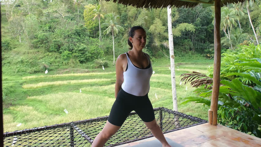 Engaged in a yoga routine, the young woman stretches gracefully on a bamboo hut terrace. Surrounded by lush tropical greenery and rice plantations, slow motion