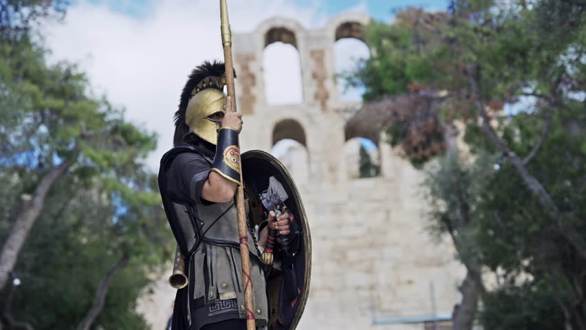 Confident hoplite spartan soldier wearing metal helmet and protecting with shield attacking with sharp spear with view of Acropolis Odeon of Herodes Atticus at background