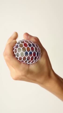 Hand squeezing a mesh stress ball with colorful beads inside against neutral background. The stress ball is compressed from different angles showing the movement of vibrant beads