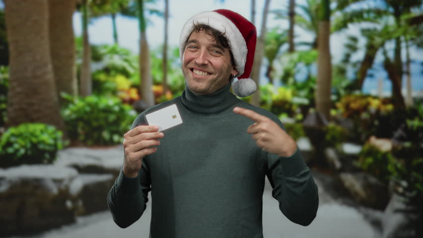 Hispanic man wearing a christmas hat joyfully points to a card outdoors on a lush street.