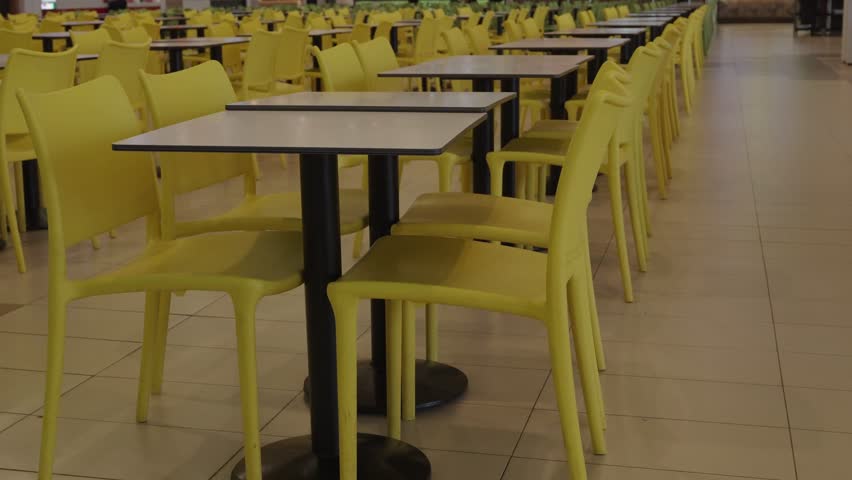 Neat rows of yellow plastic chairs and white square tables in an empty food court with tiled floor and soft lighting. Pan left