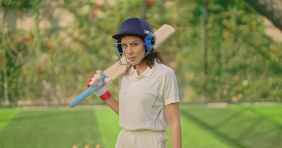 Indian gen z athlete sports woman standing at outdoor play ground wear white uniform and protective helmet looking at camera. Asian cricket player female hold bat day time at match playground