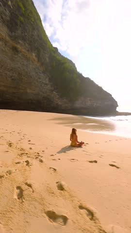 FPV drone shot of woman meditating alone on remote tropical beach with turquoise sea, waves, and cliffs, Bali, Indonesia. Peaceful scene of relaxation, mindfulness, and inner peace.