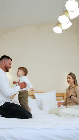 Family of three sit on the big double bed playing cheerfully. Father tosses a baby boy to mom. Fun time at home. Vertical video.
