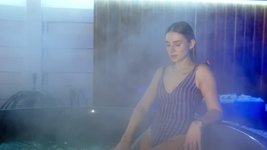 Good-looking Caucasian woman in stripy swimsuit sits on the edge of the Jacuzzi. Lady splashes water with her hand and then deeps in hot tub.