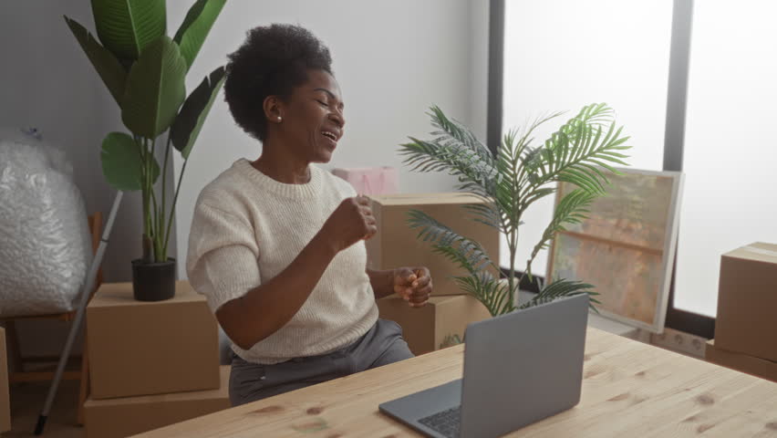 African american woman joyfully dances in her new living room surrounded by unpacked boxes and plants, celebrating with a cheerful expression.