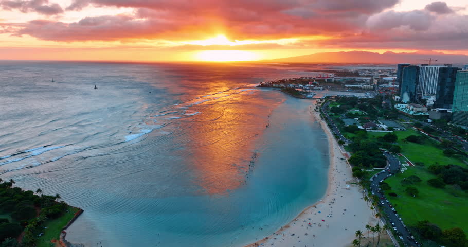 Sun sets above the Pacific Ocean. Grey clouds cover the sky above the waterscape and the shore of Honolulu, Hawaii, USA. Aerial view.