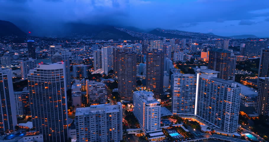 Dusk time in the modern city. Multi-storied blocks of flats switch on the lights. Thick clouds cover the mountains at backdrop. Honolulu, Hawaii, USA. Aerial view.