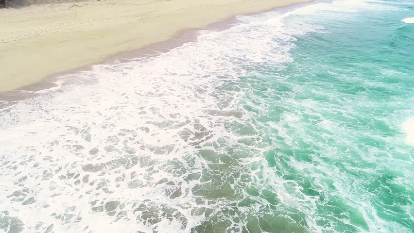 drone view aerial footage flying over a beach parallel to the water, day of waves and foam. Sea background