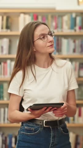 VERTICAL VIDEO: University young student preparing for exams in a modern library, searching for books with a tablet in hand. Academic or project research, self-directed learning, higher education.