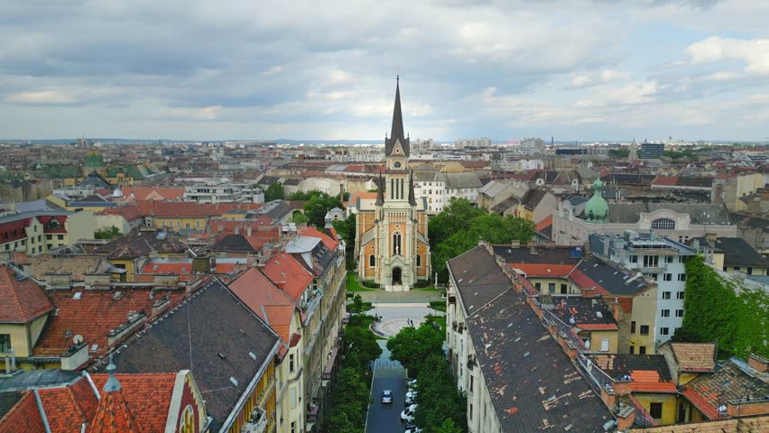 Wide aerial view of St. Francis of Assisi Parish, a neo-Romanesque Catholic church in Budapest, Hungary, surrounded by greenery and cityscape under an overcast sky.
