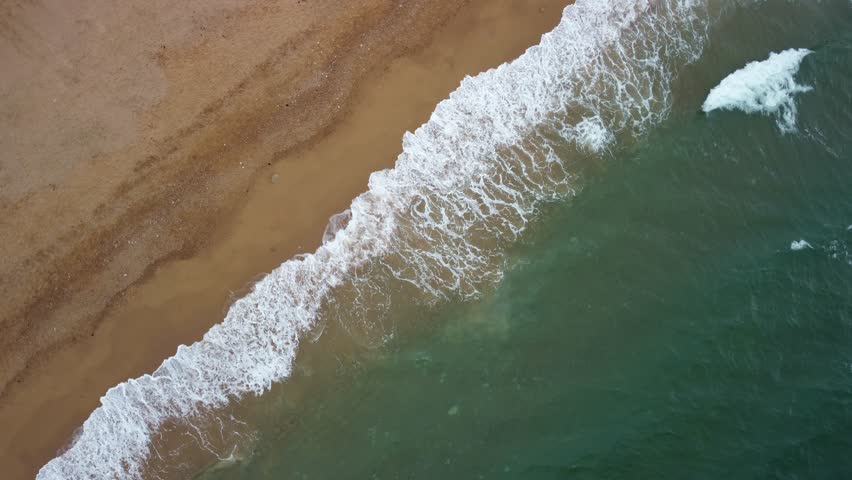 Rotating aerial footage of white waves landing on a golden sandy beach. The footage is bird eye, straight panned down angle over the waves. The drone is rotating right slowly. Jurassic Coast, Dorset.