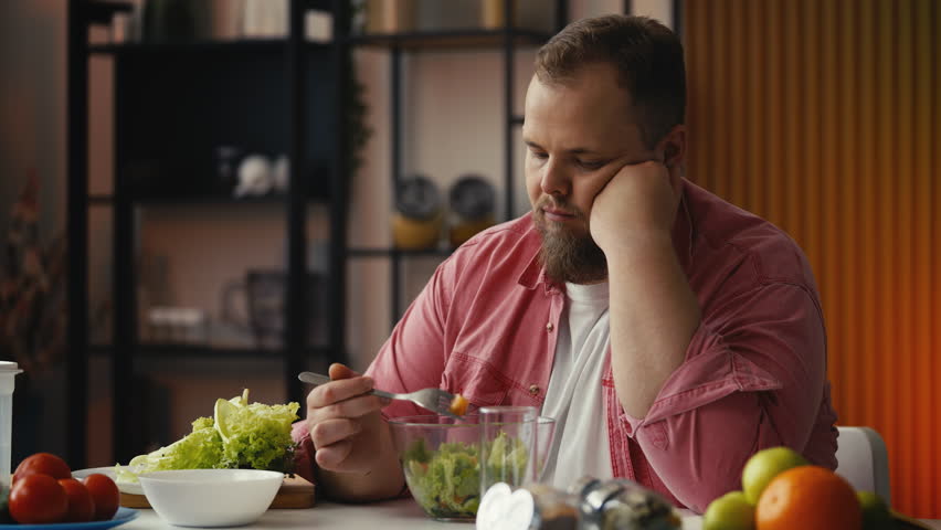 Sad plus-size man eating healthy salad with no appetite, struggling with diet