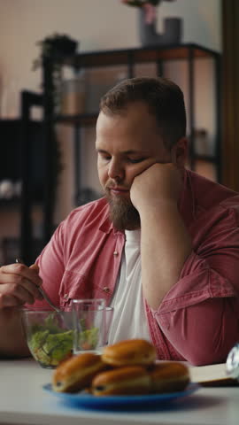 Sad plus size man eating salad while looking at pastry, struggling with cravings